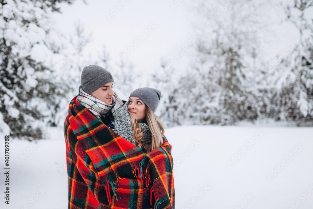 Lovers stand covered with a blanket against the background of a snowy winter forest