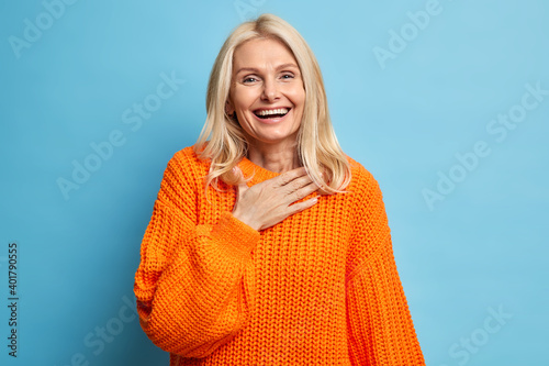 Portrait of sincere blonde woman smiles broadly has white perfect teeth keeps hand on chest feels very glad to receive help from close person wears orange knitted sweater isolated over blue background photo