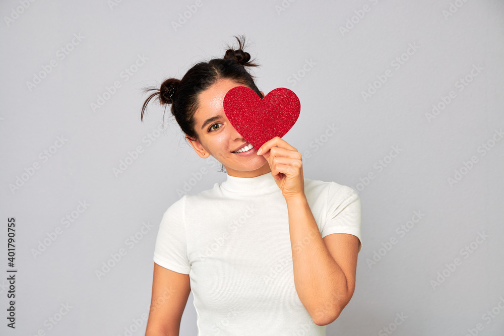 cheerful woman in love with funny hairstyle holding a small red sparkling heart valentine and covering one eye while smiling cheerful woman in love with funny hairstyle holding a small red sparkling heart valentine and covering one eye while smiling