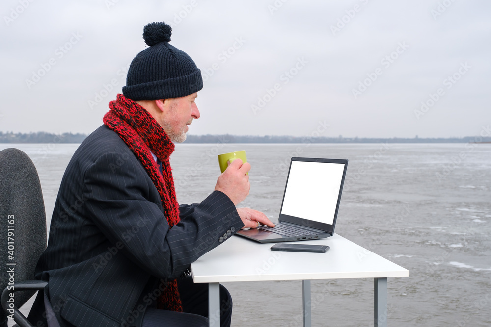 elderly bearded smiling businessman in suit looking to the camera, drinks coffee and works with laptop on table in the middle of a frozen lake. Profile view, copy space for text on device screen