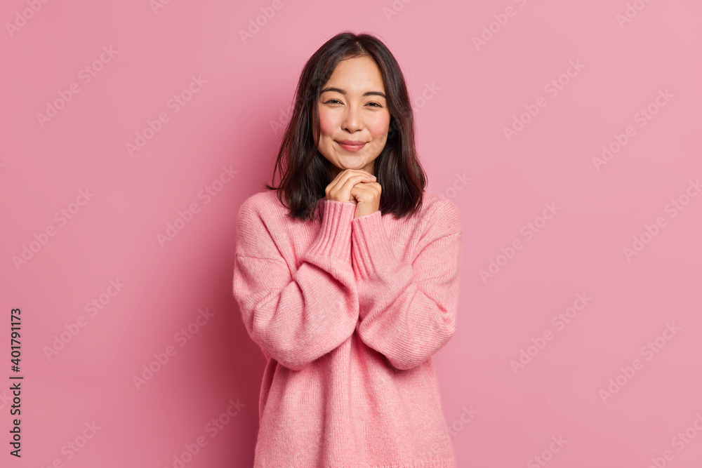 Charming brunette Asian girl with tender expression has rouge cheeks keeps hands under chin dressed in loose jumper poses against pink background. Attractive eastern woman has romantic feelings - obrazy, fototapety, plakaty 