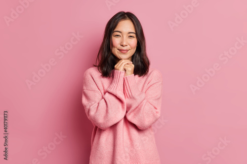 Charming brunette Asian girl with tender expression has rouge cheeks keeps hands under chin dressed in loose jumper poses against pink background. Attractive eastern woman has romantic feelings