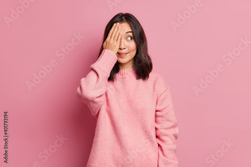 Beautiful brunette young Asian woman with eastern appearance covers eyes with hand hides face smiles pleasantly wears casual knitted sweater poses against rosy background. Dont look at me please © wayhome.studio 