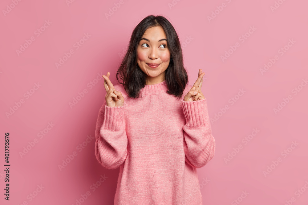 Positive brunette Asian woman crosses fingers believes in something good stands hopeful and prays for dreams come true wears casual sweater isolated over pink background. People wish desire. Positive brunette Asian woman crosses fingers believes in something good stands hopeful and prays for dreams come true wears casual sweater isolated over pink background. People wish desire.