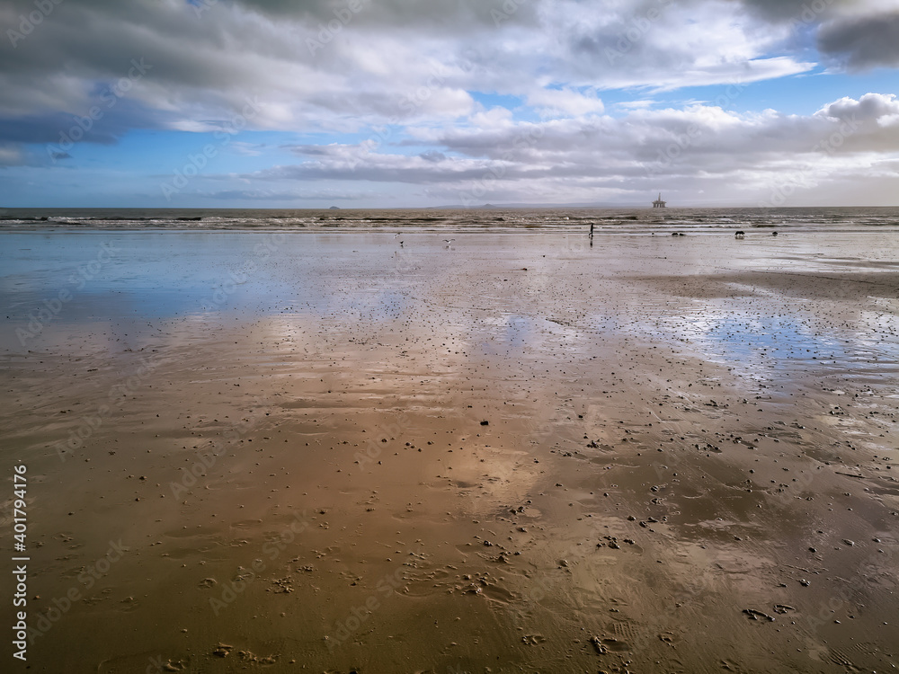 Beach at low tide with sky reflections in the water left on the sand and a dog walker and oil rigs in the distance