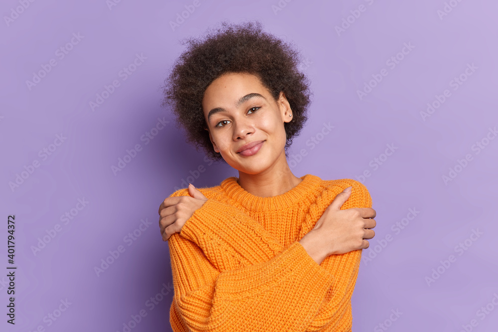 Positive Afro American teenage girl hugs herself touches shoulders expresses self love smiles pleasantly dressed in knitted orange sweater poses against purple studio background likes her new jumper Positive Afro American teenage girl hugs herself touches shoulders expresses self love smiles pleasantly dressed in knitted orange sweater poses against purple studio background likes her new jumper