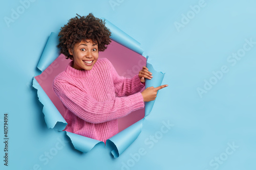 Cheerful young curly African American woman recommends something points at blank space demonstrates advertisement wears knitted pink sweater poses from blue paper hole. People promotion concept