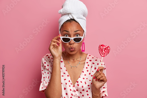 Attractive ethnic lady wears stylish sunglasses domestic silk robe and bath towel on head holds heart shaped lollipop on stick has manicure poses against pink background. Home lifestyle concept © wayhome.studio 