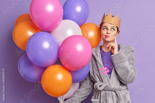 Thoughtful woman has image of queen wears crown on head stands pensive concentrated above purses lips thinks about coming holidays celebration holds multicolored inflated balloons. Holiday concept