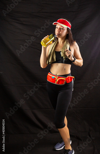 Beautiful girl in jogging red uniform, drinks water after training in the studio on a black background
