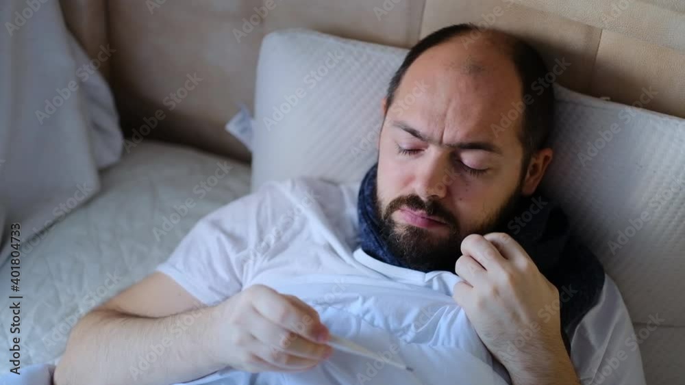 The man is sick in bed, checking the temperature. A person having a high fever watching a thermometer. The man is sick at home. The man is sick in bed, checking the temperature. A person having a high fever watching a thermometer. The man is sick at home.