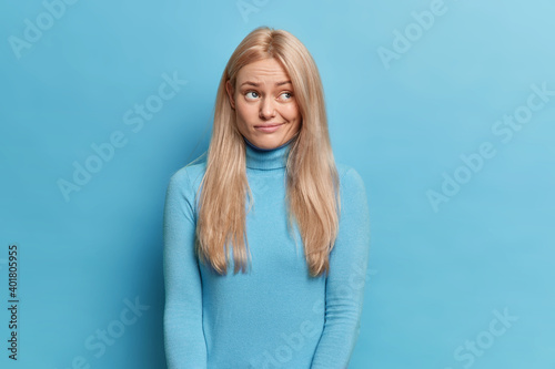 Displeased blonde young woman with long hair looks upset aside has thoughtful expression purses lips wears turtleneck isolated over blue background. Human face expressions and emotions concept