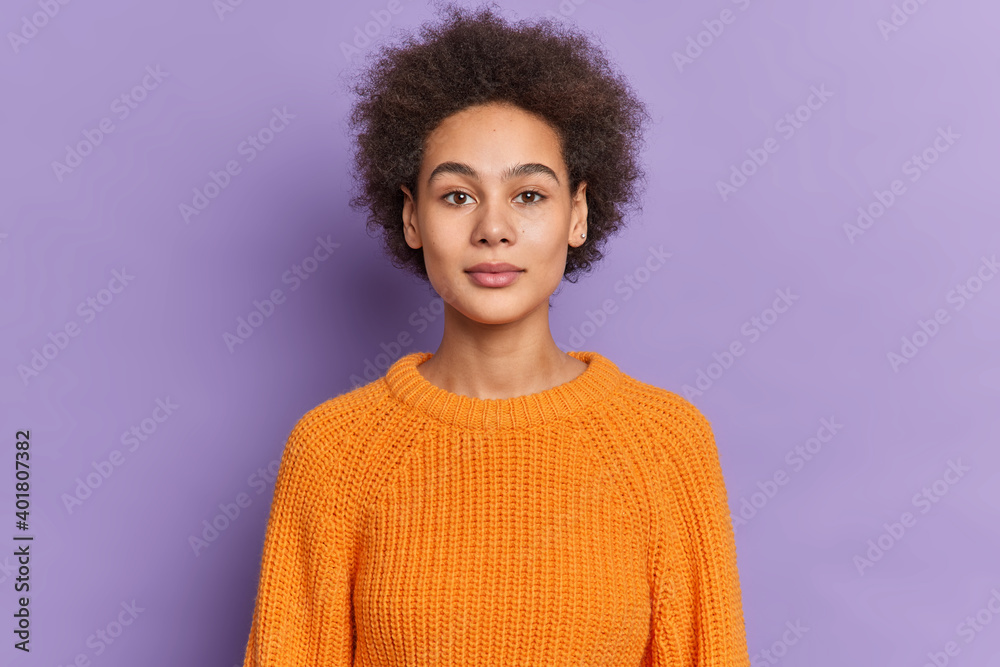 Studio shot of serious dark skinned female teenager with curly bushy hair looks confidently at camera has natural beauty calm expression dressed in knitted sweater isolated over purple background Studio shot of serious dark skinned female teenager with curly bushy hair looks confidently at camera has natural beauty calm expression dressed in knitted sweater isolated over purple background