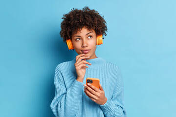 Photo of beautiful African American woman listens music with smartphone and headpones has thoughtful expression dressed in knitted jumpper isolated over blue background. Quarantine leisure concept
