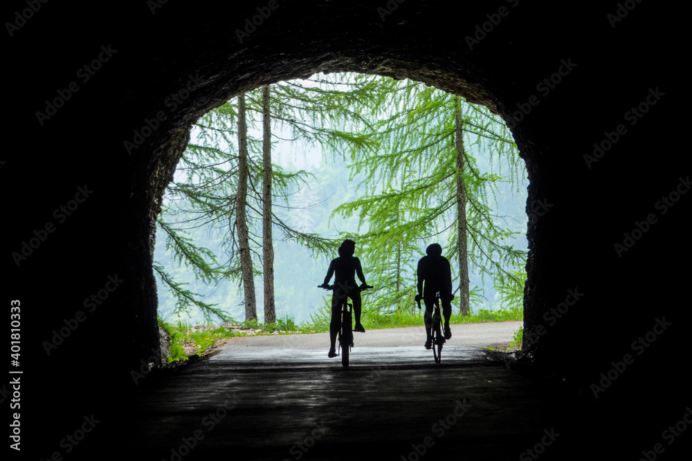 Two unrecognizable tourists ride an electric and road bicycle out of a tunnel.