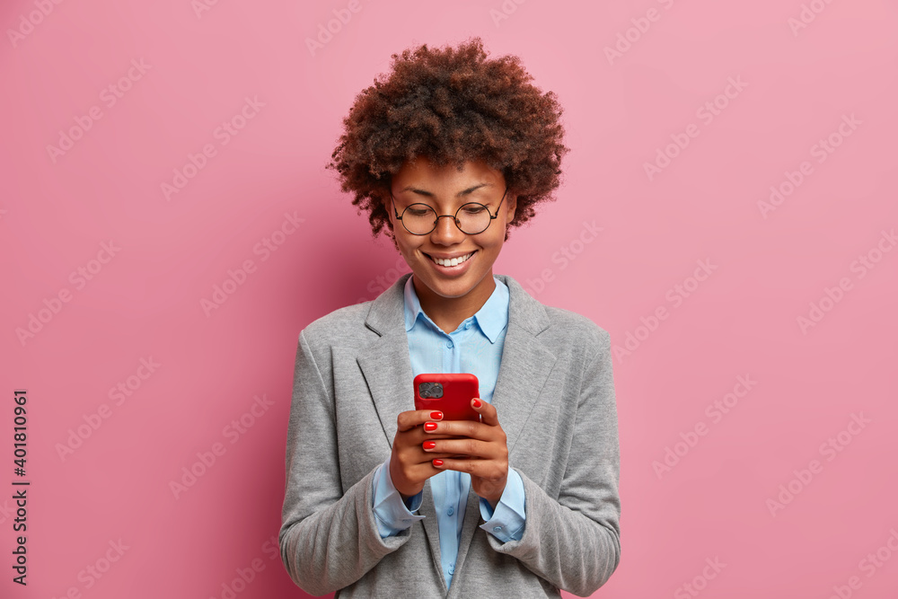 Horizontal shot of glad dark skinned successful woman dressed in formal wear uses mobile phone sends text messages reads business news isolated over pink background. People and technology concept - obrazy, fototapety, plakaty 