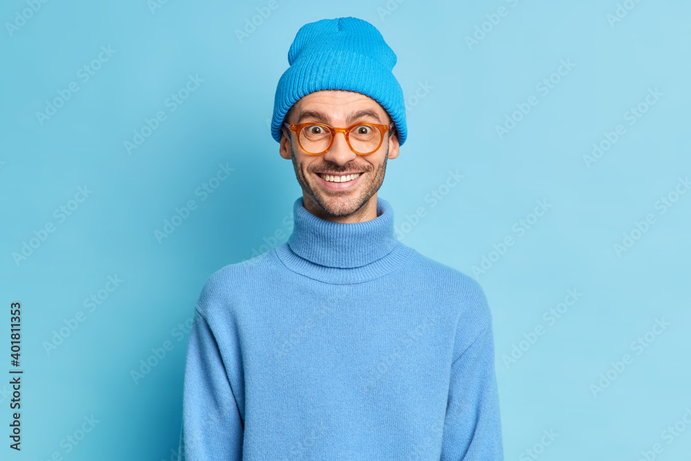 Studio shot of handsome cheerful guy has positive face expression looks through optical glasses wears hat and turtleneck isolated over blue background. Male teenager in stylish clothes poses indoor