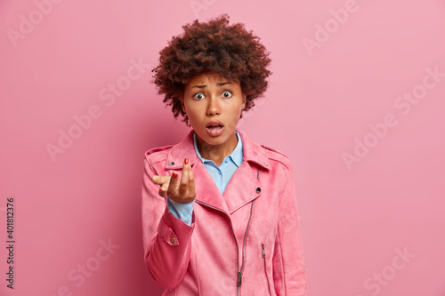 Frustrated puzzled African American woman raises palm and looks indignant at camera keeps mouth opened reacts on hearing something bad dressed in stylish jacket isolated over pink background