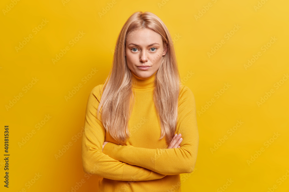 Obraz premium Studio shot of self confident blonde young woman keeps arms folded looks seriously at camera dressed in casual turtleneck isolated over yellow background. Pretty female model in assertive pose
