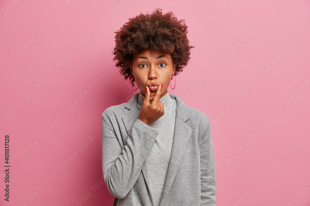 Photo of dark skinned woman keeps lips folded makes grimace looks directly at camera has curly bushy hair dressed in elegant grey clothing isolated over pink background. Human face expressions Photo of dark skinned woman keeps lips folded makes grimace looks directly at camera has curly bushy hair dressed in elegant grey clothing isolated over pink background. Human face expressions