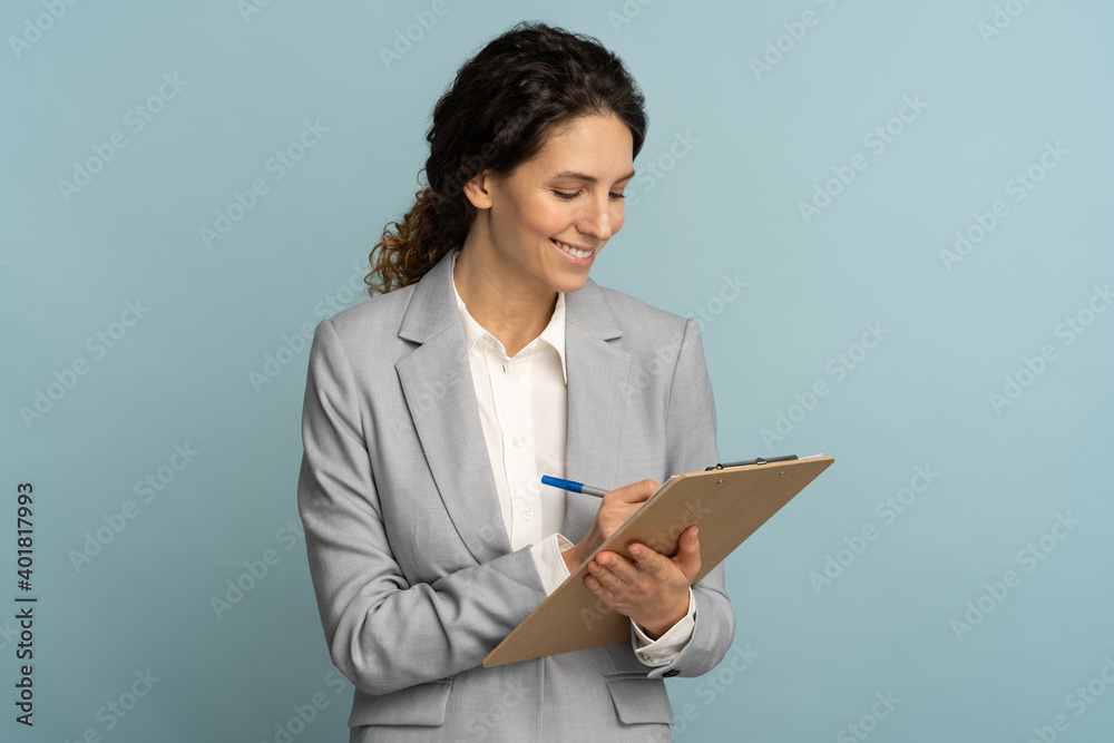 Teacher woman wear grey blazer and white blouse writing, makes notes in document, holds folder in her hand. Female office worker isolated on studio blue background. Teacher woman wear grey blazer and white blouse writing, makes notes in document, holds folder in her hand. Female office worker isolated on studio blue background.