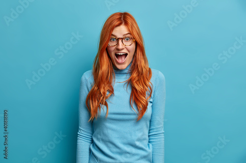 Young redhead woman with shocked expression keeps mouth opened reacts in astonishing news wears turtleneck isolated over blue background. Pretty foxy girl being greatly surprised by something