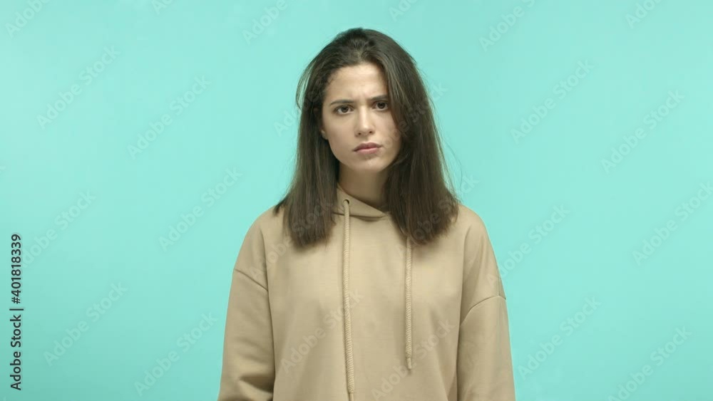 Angry young woman in beige hoodie staring annoyed and showing hush sign, telling to shut up, be quiet gesture, standing over turquoise background Angry young woman in beige hoodie staring annoyed and showing hush sign, telling to shut up, be quiet gesture, standing over turquoise background