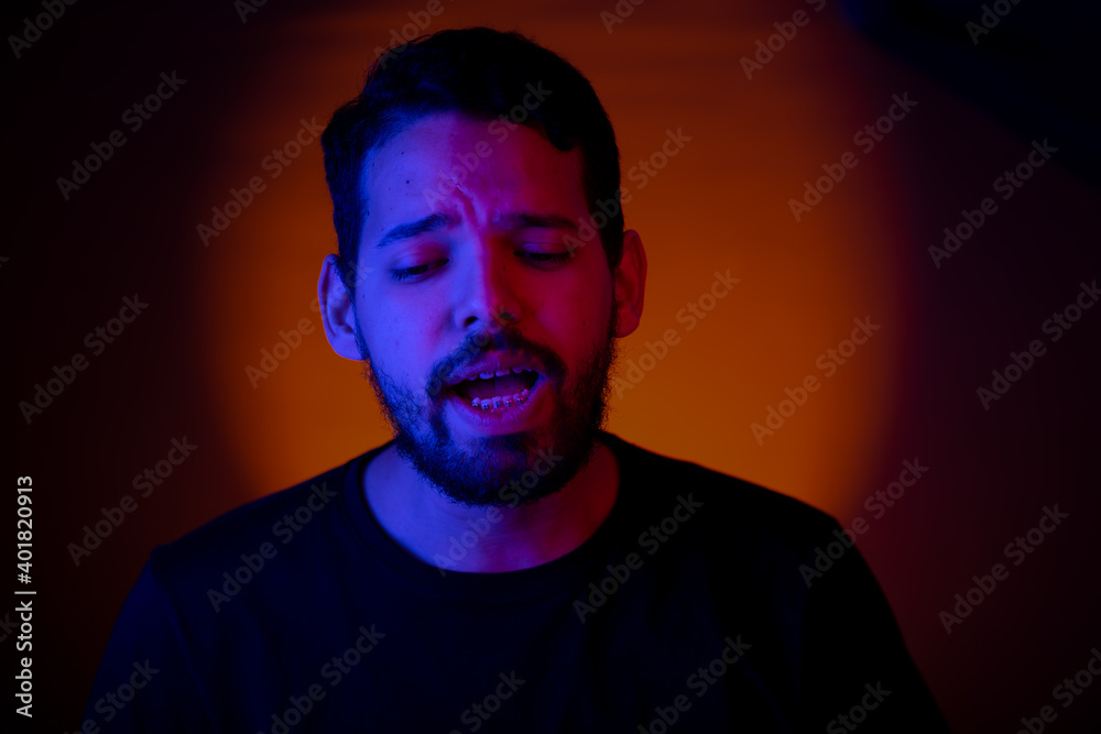 Fototapeta premium Young man portrait with colored lights in a studio shot with an orange light in the background. He is singing along with the music in his party.
