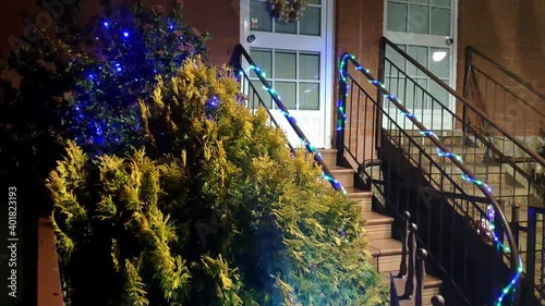 Entrance of a house decorated for Christmas. The front door has a wreath and there is a tree decorated with blue flashing lights. On the front steps handrail there is a ornamental lights strip. photo