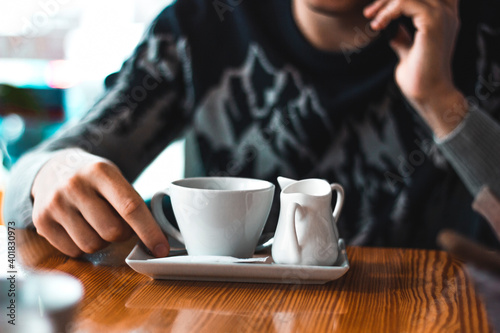 Man talking over the phone in a cafe with a coffee cup and cream milk pot on the table. Adult hand and winter sweater