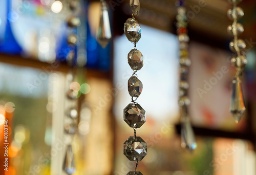 String with chandelier crystals catching light. In the background other crystal strings, glass flasks and other objects. A detail of a window full with glass and other translucent objects.