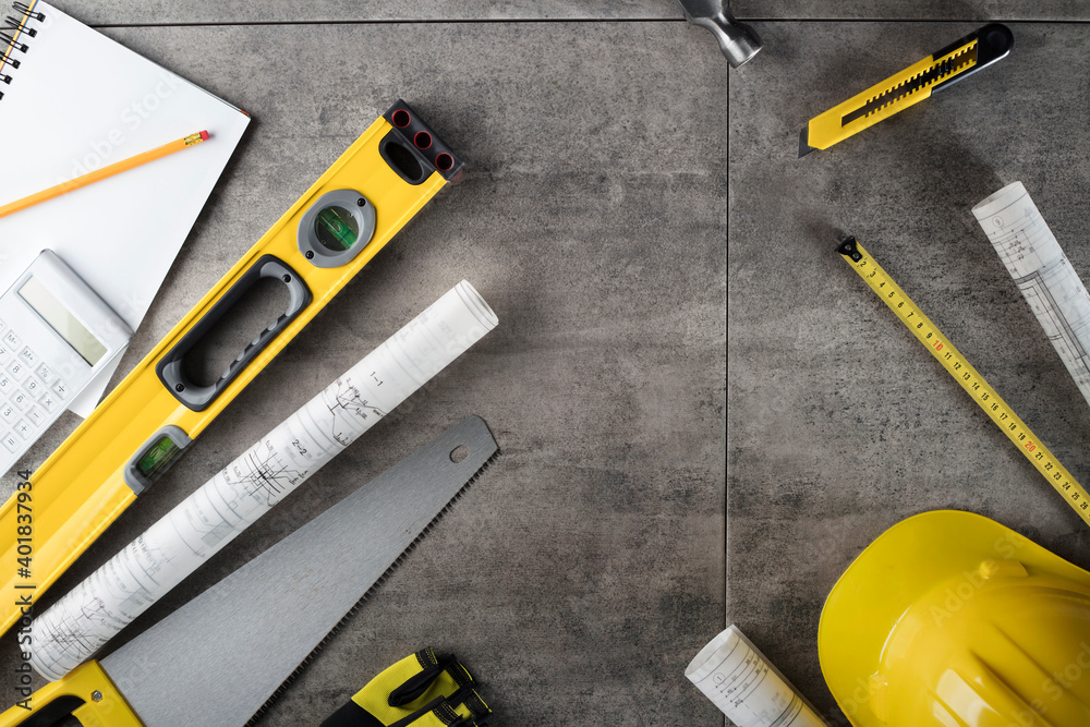 Contractor concept. Tool kit of the contractor: yellow hardhat, libella, hand saw. Plans and notebook on the gray tiles background. - obrazy, fototapety, plakaty 