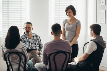 Middle age woman stands in front of a group of people during a therapy meeting and talks about her family problems