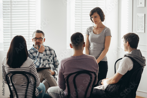 Middle age woman stands in front of a group of people during a therapy meeting and talks about her family problems © Photographee.eu