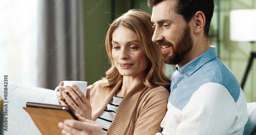 Obraz premium Portrait of young Caucasian joyful couple shopping on internet browsing online on tablet device sitting on sofa in apartment spending time together with cup of tea. Husband and wife resting at home
