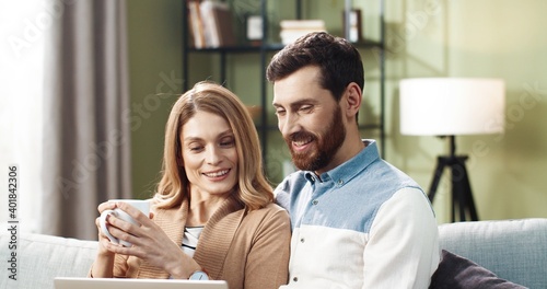 Portrait of young Caucasian joyful couple shopping on internet browsing online on tablet device sitting on sofa in apartment spending time together with cup of tea. Husband and wife resting at home