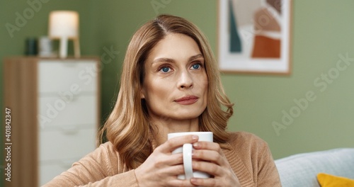 Close up of cheerful young Caucasian pretty woman in positive mood resting relaxing on sofa in cozy home sipping hot drink from cup. Drinking coffee and smiling at camera. Leisure concept