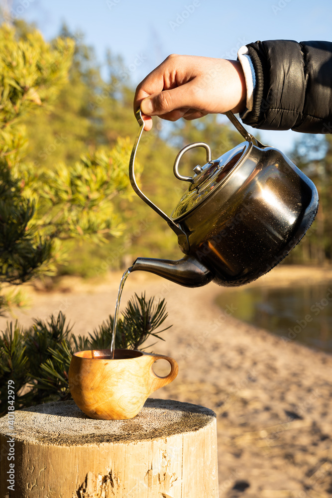 A man pours hot coffee or tea from a vintage teapot into a wooden cup on a stump.

