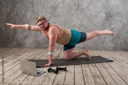 funny fat man in sports clothes doing exercises. He plays sports remotely while looking at his laptop