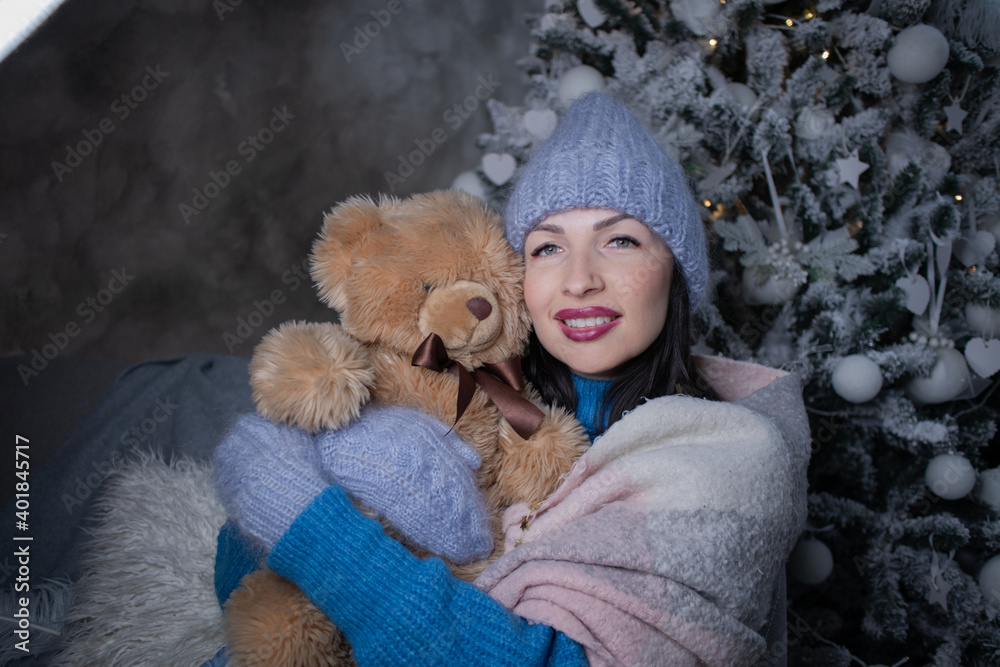 Fototapeta premium brunette in a blue sweater and blue hat and mittens with a teddy bear near the new year tree