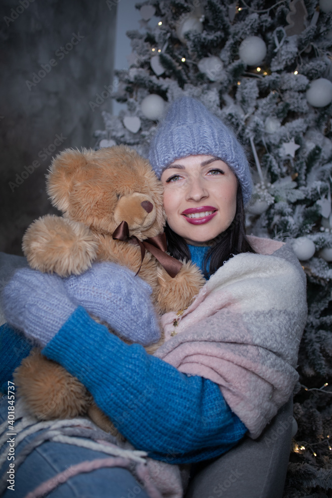 Fototapeta premium brunette in a blue sweater and blue hat and mittens with a teddy bear near the new year tree