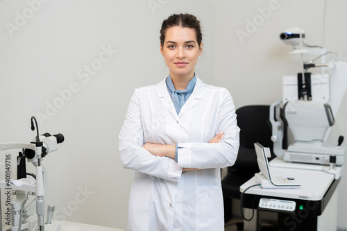 Young smiling female ophthalmologist in whitecoat crossing her arms by chest