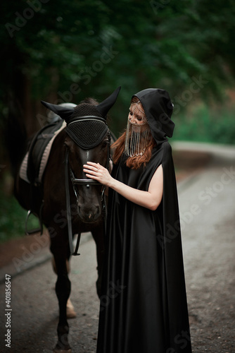 Young woman in hat and black cloak with a horse in the forest