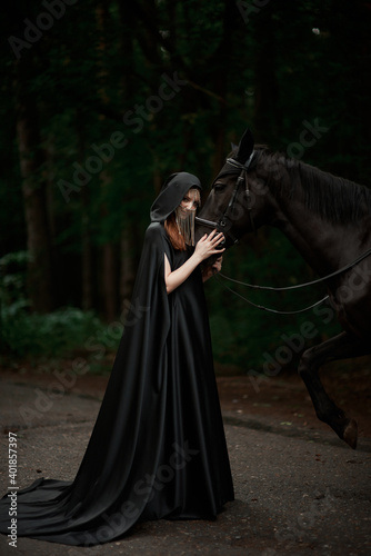 Young woman in hat and black cloak with a horse in the forest