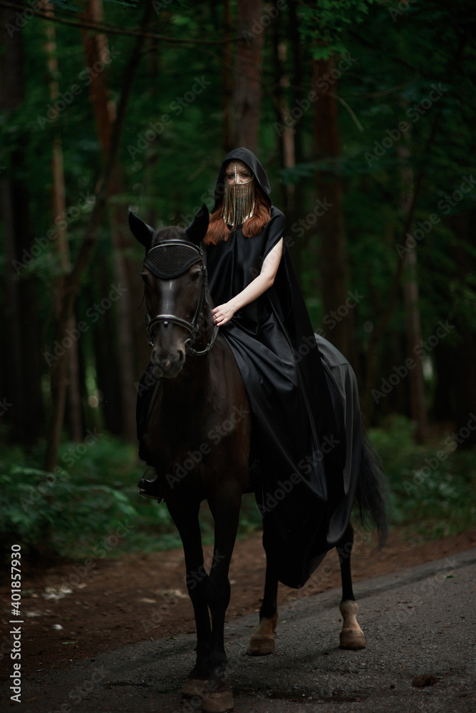 Young woman in hat and black cloak with a horse in the forest Young woman in hat and black cloak with a horse in the forest