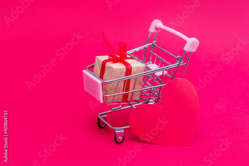 Small grocery cart with gift boxes on red-pink background. Give gifts with love on Valentine's Day, Christmas and birthday. Shopping online. Holiday sales and discounts. Retail and wholesale purchase