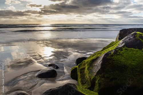 The setting sun grazes moss covered boulders as the sky reflects on the wet sand in Carlsbad, CA.