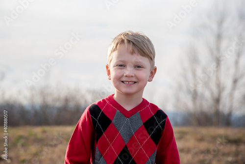 Outdoor winter photo of an adorable happy young boy with blond hair smiling, wearing a red sweater and looking at the camera