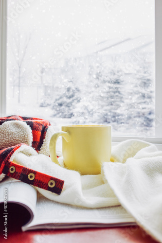 Yellow mug of hot tea with a white woolen scarf and classic plaid pattern warm hat put on an open book at the window with landscape view of snow falling on pine trees in winter season. Cozy living.