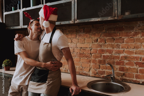 Loving gay couple at home. Two handsome men hugging and kissing on kitchen in Santa Claus hat. Christmas mood. LGBT concept.